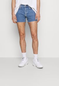 Ljust blå denimshorts med fransade kanter, bärs tillsammans med vita sportskor och ankelstrumpor. Enkel, avslappnad design med standardfickor.