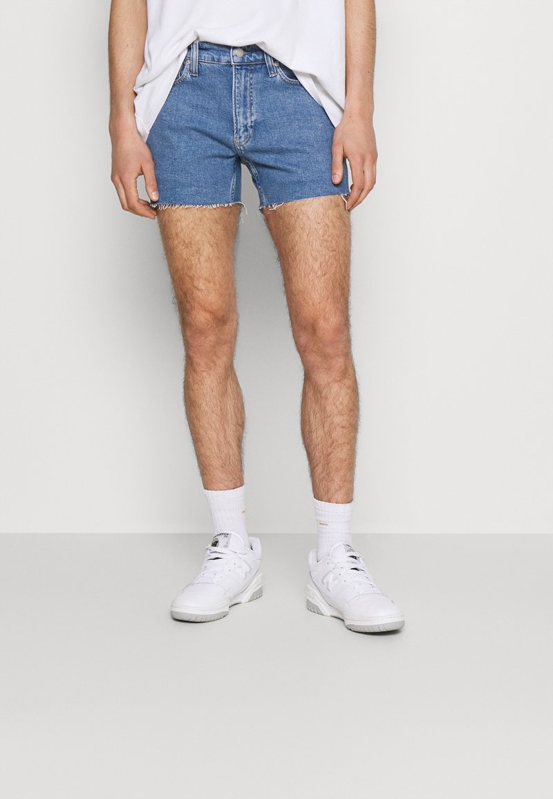 Ljust blå denimshorts med fransade kanter, bärs tillsammans med vita sportskor och ankelstrumpor. Enkel, avslappnad design med standardfickor.