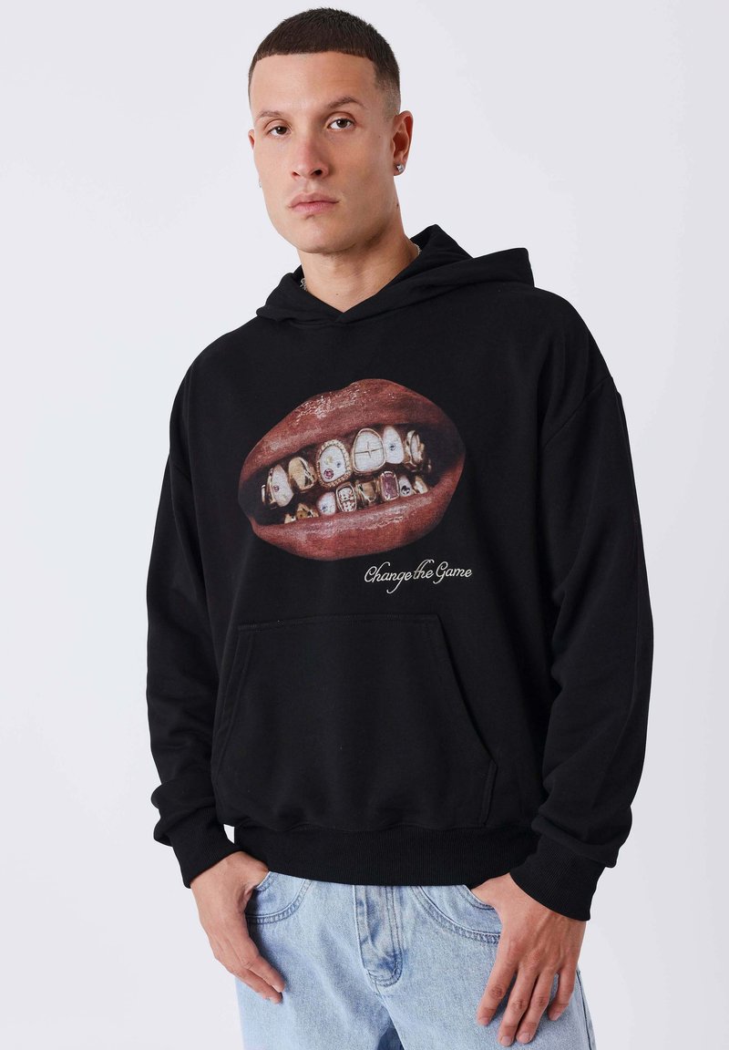Homme portant un sweat à capuche noir avec un motif de lèvres montrant des dents en or et ornées de bijoux, accompagné du texte « Change the Game », et un jean bleu clair.