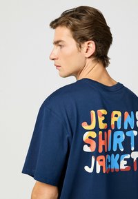 Marinblå t-shirt med kort ärm och en texturerad, färgglad grafik med texten "JEANS SKJORTA JACKA" på baksidan, med djärva, lekfulla typsnitt.