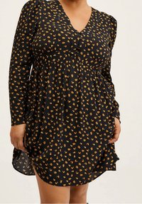 Abito nero con scollatura a V, maniche lunghe e vita elastica. Presenta un piccolo motivo floreale giallo e una silhouette leggermente svasata.