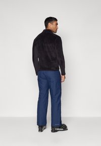 Schwarze Fleecejacke aus strukturierter stoff mit hohem Kragen, kombiniert mit blauen Skihosen und schwarzen Stiefeln mit grünen Akzenten.