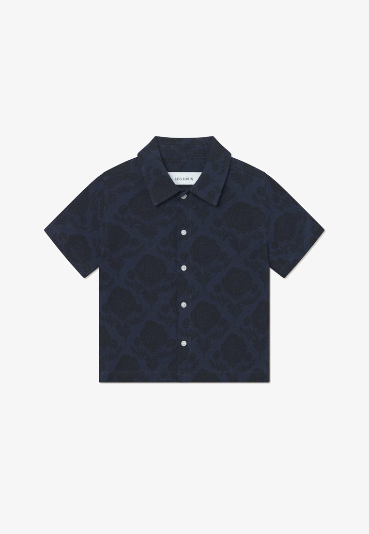 Μπλούζα κοντομάνικη από ναυτικό μπλε ύφασμα με λουλουδάτο σχέδιο, διαθέτει κολάρο με κουμπιά και μπροστινό παρμπρίζ. Τα κουμπιά είναι λευκά.