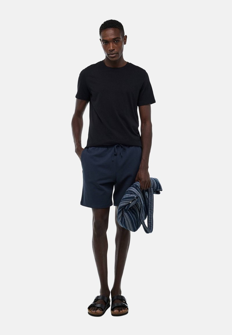 T-shirt noire, shorts navy avec cordon de serrage, et sandales noires. Tenant une serviette bleue rayée. Design simple, coupe décontractée, look décontracté.