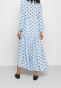 Femme portant une robe maxi à manches longues de couleur bleu clair avec des pois noirs, se tenant de dos face à la caméra et chaussée de noir.