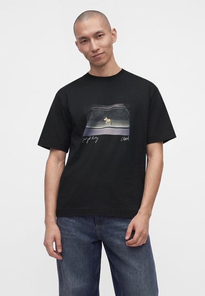 Homme portant un t-shirt noir avec un petit dessin de lapin jaune et du texte, associé à un jean bleu, debout devant un fond blanc uni.