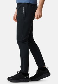 Zwarte sportbroek met een slim fit, gemaakt van lichtgewicht materiaal, met zijzakken en ritsen bij de enkels. Gecombineerd met zwarte sneakers.