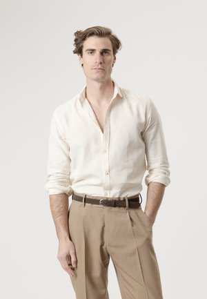Hombre con cabello castaño claro que lleva una camisa beige abotonada con las mangas remangadas y pantalones marrón claro con un cinturón marrón oscuro, de pie frente a un fondo simple.