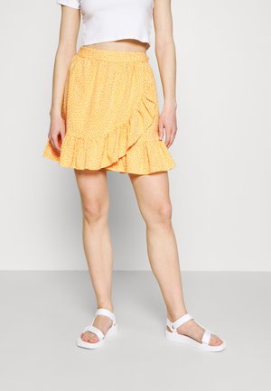 Kvinde iført hvid crop top, gul flæset nederdel med hvide prikker og hvide åbne sandaler står mod en ensfarvet baggrund.