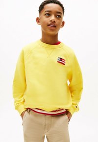 Felpa gialla con polsini e scollatura a costine, con un piccolo patch logo multicolore. Indossata sopra una camicia a righe rossa e bianca.