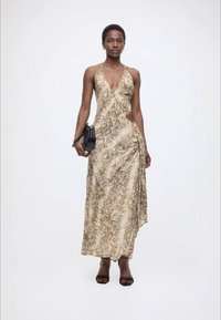 Γυναίκα που φοράει μπεζ αμάνικο φόρεμα με μοτίβο φίδι και βαθύ ντεκολτέ, κρατώντας μαύρη τσάντα και φοράει μαύρες ψηλοτάκουνες γόβες, στέκεται μπροστά από λευκό φόντο.