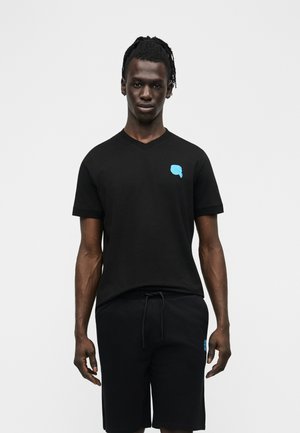 Dorosły mężczyzna ubrany w czarny T-shirt z dekoltem w serek i czarne szorty, oba z małym niebieskim nadrukiem po lewej stronie, stojący na białym tle.