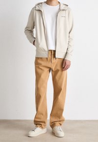 Felpa con zip beige dal design minimalista, abbinata a pantaloni larghi marrone chiaro e sneakers bianche. Tessuto morbido e stile casual.