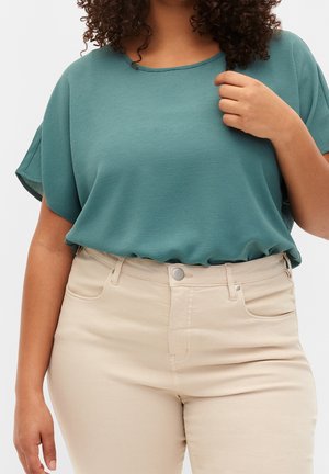 Woman wearing teal short-sleeve blouse tucked into high-waisted beige pants, with one hand touching her collar.