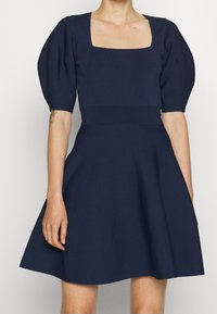 Tmavě modré šaty se čtvercovým výstřihem, nabíranými rukávy a vypasovaným pasem. Sukně se rozšiřuje, vyrobená z hladkého texturovaného materiálu.
