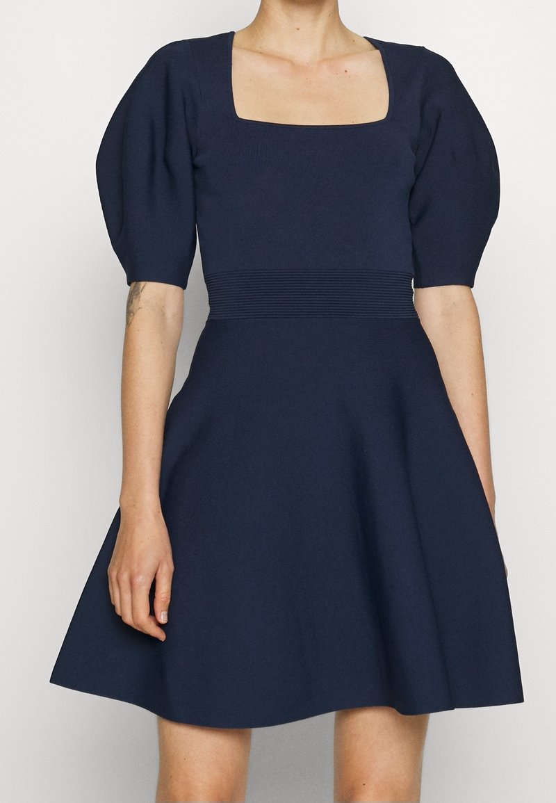 Tmavě modré šaty se čtvercovým výstřihem, nabíranými rukávy a vypasovaným pasem. Sukně se rozšiřuje, vyrobená z hladkého texturovaného materiálu.