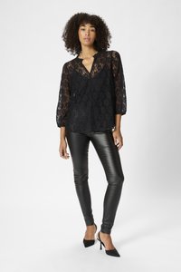 Schwarze Spitzenbluse mit Dreiviertelärmeln, tiefem V-Ausschnitt und gewellten Kanten, kombiniert mit enganliegenden schwarzen Lederhosen und schwarzen High Heels.