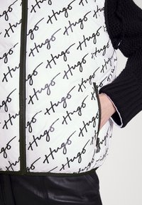 Prošívaná vesta z bílé tkaniny s opakujícím se černým vzorem loga „Hugo“. Má přední zip a olivově zelený lem.