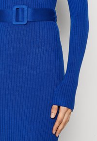 Gros plan d'une personne portant une robe ajustée, côtelée, bleu vif avec une ceinture assortie et des manches longues dépassant le poignet.
