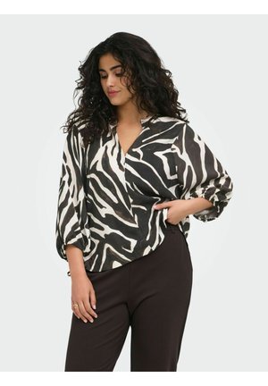 Kvinde med krøllet hår iført en sort-hvid zebra-stribet bluse med trekvart puffede ærmer og sorte bukser, stående med den ene hånd i lommen.