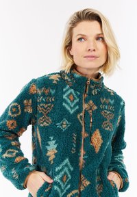 Žena s krátkymi blond vlasmi, oblečená v teal, oranžovej a béžovej vzorovanej fleecovej bunde so zipsom, má ruky v vreckách a pozerá sa mierne na bok.