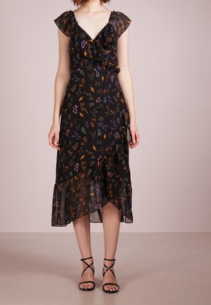 Frau trägt ein schwarzes Midikleid mit Blumenmuster, gerafftem V-Ausschnitt und asymmetrischem Saum, kombiniert mit schwarzen, Riemchen-Sandaletten mit hohen Absätzen.