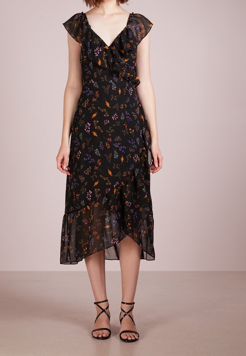 Femme portant une robe midi noire à fleurs avec un col en V volanté et un ourlet asymétrique, associée à des sandales noires à talons hauts et fines brides.