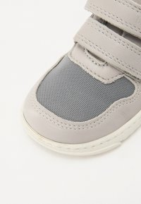 Pelēki sporta apavi ar gludu ādas purngalu, sietiņu vidusdaļu un diviem Velcro siksniņām. Balta gumijas zole ar nedaudz redzamām nolietojuma pazīmēm.