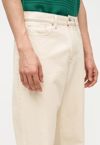 Κοντινό πλάνο ενός ατόμου που φοράει εκρού τζιν και πράσινο πλεκτό τοπ, με τα χέρια χαλαρά στα πλευρά τους.