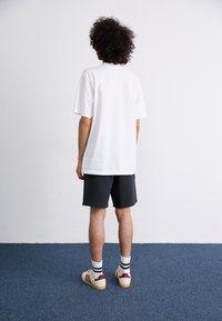 Bílý bavlněný tričko s volným střihem, spárované s tmavými šortkami. Béžové tenisky s vínovými akcenty, pruhované ponožky. Jednoduché pozadí, minimální textura.