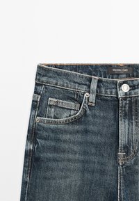 Jean en denim bleu avec poche avant, poche pour pièce de monnaie, passants de ceinture et fermeture par bouton métallique, étiqueté Massimo Dutti, fabriqué en Turquie.
