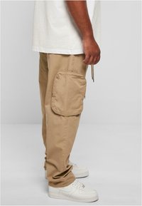 Khaki cargo pants with a large side pocket, made of cotton. Fitted with a drawstring waist, paired with white sneakers.