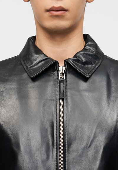 Άνδρας που φοράει μαύρο δερμάτινο μπουφάν με ασημένιο φερμουάρ, με εμφανές το γιακά, mostrando λαιμό και κάτω μέρος προσώπου μπροστά σε ουδέτερο φόντο.
