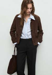 Chaqueta de ante marrón con cuello clásico, dos bolsillos frontales y cierre de botones, combinada con una camisa a rayas azul clara y pantalones negros.