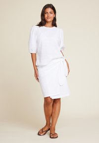 Hvit linbluse med pufferm, kombinert med en belted hvit linbunn. Modell iført brune flip-flops står mot en nøytral bakgrunn.
