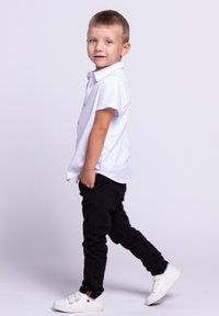 Biała koszula z krótkim rękawem z guzikami, czarne spodnie i białe trampki. Pozowanie w stojącej pozycji ukazuje casualowy ubiór i lekki materiał.