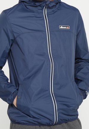 Granatowa kurtka na suwak z kapturem, biała linia zamka błyskawicznego, przednie kieszenie i logo Ellesse na piersi.