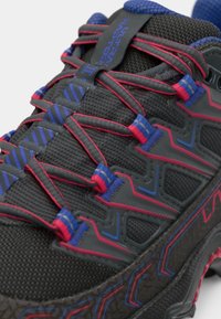 Zapatilla deportiva negra con parte superior texturada, que presenta acentos rojos y azules en los cordones y alrededor de los laterales. Marca notable en la lengüeta.