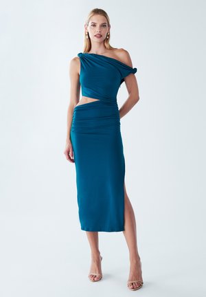 Teal silkinen mekko, jossa on yksi olkapää design, sivuleikkaus ja kerrostettu tekstuuri. Keskipituinen malli, jossa on korkea halkio ja neutraalit korot.