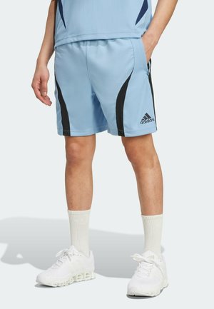 Osoba nosząca jasnoniebieskie szorty Adidas z czarnymi paskami, białe skarpetki i białe buty Adidas, stojąca z jedną ręką w kieszeni.