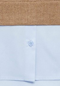 Gros plan sur l’ourlet d’une chemise bleu clair boutonnée sous le bord d’un pull en tricot côtelé marron.