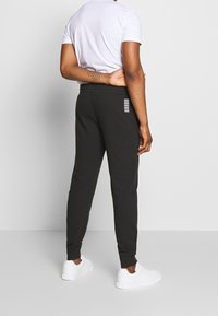 Svarta joggers med tapered fit, elastisk midja och subtila vita ränder på bakfickan. Modellen bär vita sneakers och en grå T-shirt.