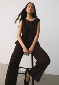 Žena s dlouhými tmavými vlasy, oblečená v tmavé bezrukávové vestě a širokých kalhotách, sedící na stoličce před jednoduchým pozadím.