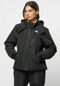 Schwarze wasserdichte Jacke mit hohem Kragen, verstellbare Kapuze, Reißverschlusstaschen und Klettverschluss-Akzenten. Mit einem Logo auf der linken Brust.