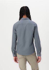 Camisa gris de manga larga hecha de tela suave, con collar clásico, puños abotonados y un dobladillo curvado, vista desde la parte de atrás.