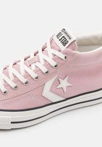 Light pink canvas sneaker with white laces and rubber sole. Features a white star logo on the side and contrasting black stripe on the sole.