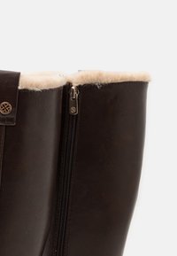 Gros plan d'une botte en cuir brun foncé avec doublure en fourrure beige et fermeture éclair latérale avec tirette en métal décorative.