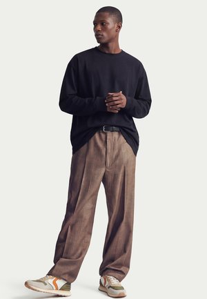 Uomo in piedi con le mani intrecciate, indossa una maglia nera a maniche lunghe, pantaloni marroni a quadri a gamba larga, cintura nera e scarpe da ginnastica bianche con accenti arancioni e verdi.