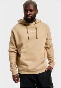 Beige hette med frontlomme, snorhette, og hvit brodert logo. Mykt, bomullsbåndmateriale med ribbede mansjetter og kant.