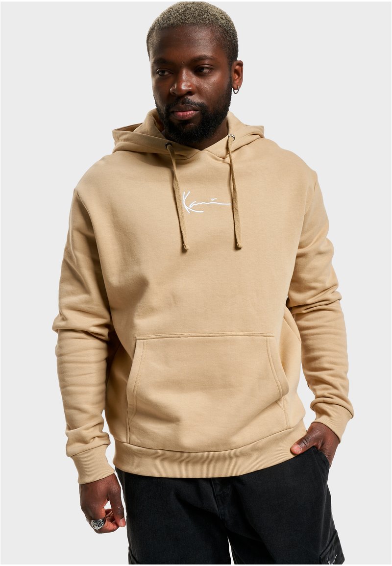 Beige hette med frontlomme, snorhette, og hvit brodert logo. Mykt, bomullsbåndmateriale med ribbede mansjetter og kant.